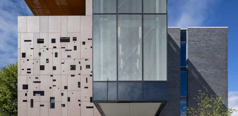 Image of a tall modern set of buildings, one brown with cut out windows one grey and pointed, with steps up beside a tree