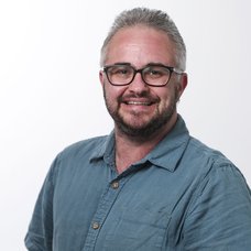 A grey-haired white man with brown and grey beard and brown-framed glasses, wearing a blue-green shirt.